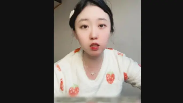 Una joven china llamada Li Yixue mira a la cámara, vistiendo una blusa blanca con fresas decorándola, lápiz labial rojo y el cabello atado hacia atrás y sujeto con un pasador.