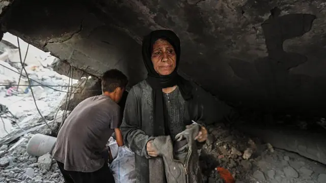 Una mujer con un pañuelo oscuro en la cabeza y un vestido gris sostiene un jersey en sus manos mientras revisa los escombros encontrados en un mercado de Gaza que fue atacado por Israel. Detrás de ella hay un niño de espaldas a la cámara.