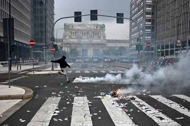 Un manifestante de espaldas hace el gesto de dar una patada en medio de una calle de Milán, con humo a su derecha y vehículos de la policía antidisturbios al fondo, cerca de la estación de tren Milano Centrale.
