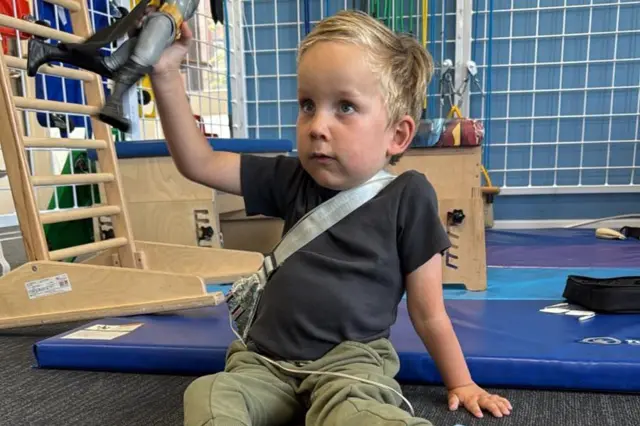 Edward está sentado en el piso sosteniendo un muñeco de juguete. Viste una playera oscura y pantalón de sudadera verde 