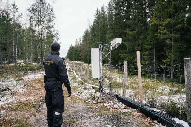 Un militar uniformado mira a lo largo de una larga verja con postes de madera que divide un territorio boscoso con parches de nieve