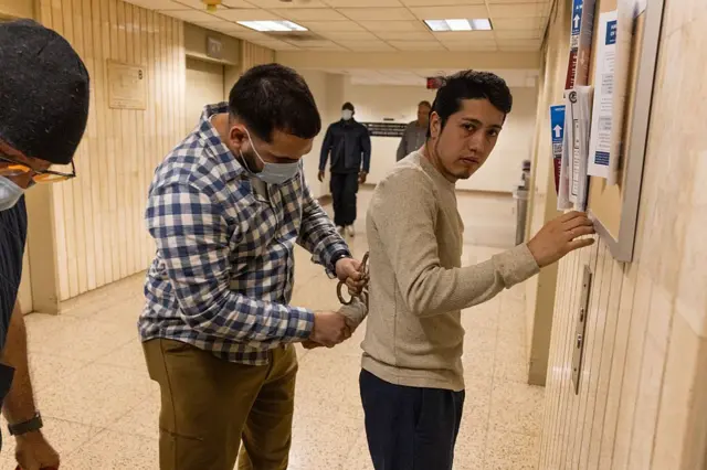 Un agente de paisano de ICE con el rostro cubierto esposa a un joven inmigrante en un tribunal de Inmigración de Nueva York.