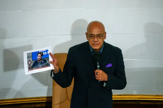 El presidente de la Asamblea Nacional de Venezuela, Jorge Rodríguez, vestido con un traje oscuro sostiene una fotografía de la primera ministra de Trinidad y Tobago.