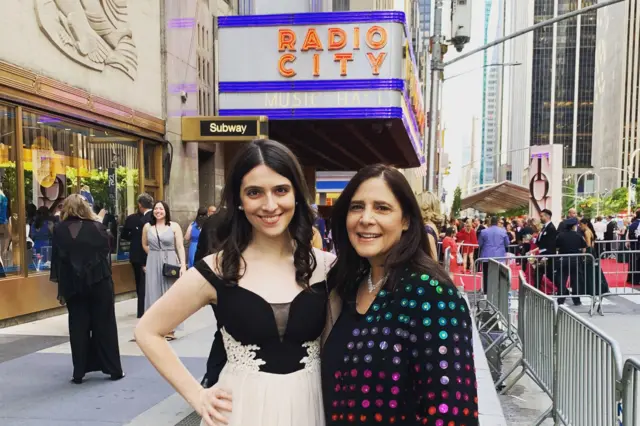 Sammi y su madre posan para una fotografía delante del Radio City Music Hall de Nueva York. Sammi lleva un elegante vestido blanco y negro y su madre lleva una chaqueta. Detrás de ellas hay otras personas que también posan para las fotografías. Una alfombra roja está formada por círculos multicolores.