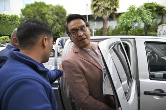 Fernando Villavicencio, con chaqueta y gafas y medio cuerp, habla con dos personas vestidas de azul, junto a una camioneta con la puerta abierta a la que parece estar entrando. 