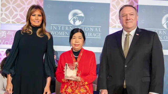 Susanna Liew lleva una elaborada chaqueta bordada en rojo y amarillo y sostiene un trofeo de cristal, sonriendo levemente, mientras permanece en el escenario entre Melania Trump, que lleva un vestido negro, y Mike Pompeo, que lleva un traje negro y corbata dorada.