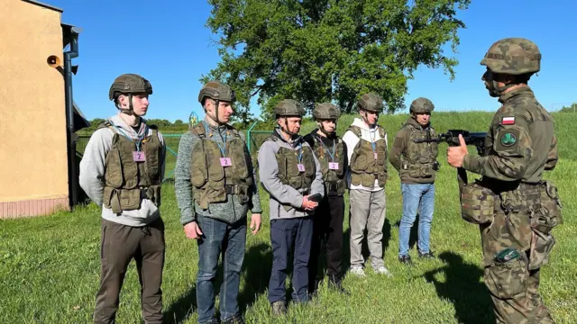 Una fila de seis personas con cascos y chalecos de camuflaje observa a un soldado con uniforme de camuflaje y una insignia de la bandera polaca que les muestra un arma. Están de pie en un campo.