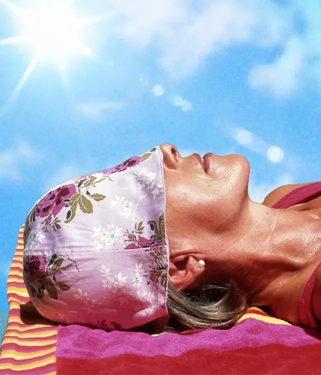 Una mujer recostada tomando el sol con un gorro morado, con flores moradas, que le cubren los ojos.
