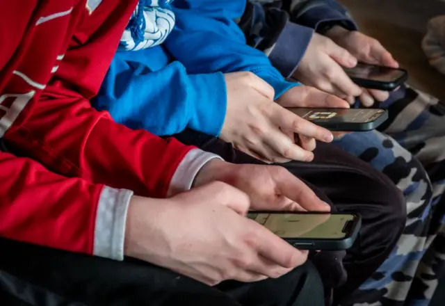 Fotografía de tres chicos a los que no se les ve el rostro pero que están sentados sosteniendo sus teléfonos celulares en sus manos.