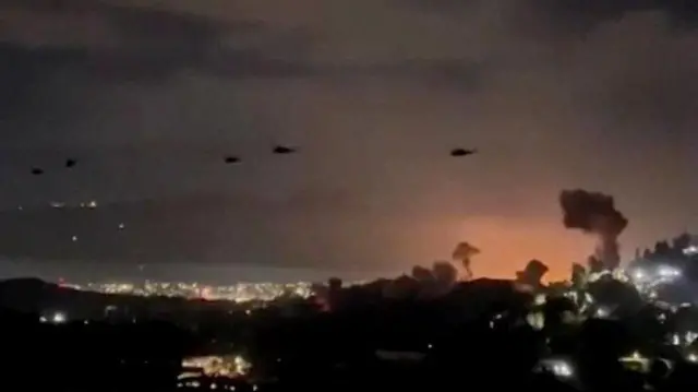 Imagen fija ligeramente borrosa de un video que muestra al menos 5 helicópteros volando sobre Caracas, y se pueden ver columnas de humo que emanan del suelo.