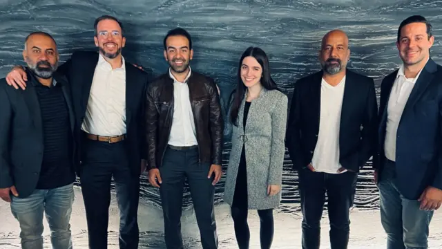 Safi y Sammi sonriendo mientras están de pie con dos hombres a cada lado. Uno de los hombres tiene el brazo sobre los hombros del hombre que tiene al lado.