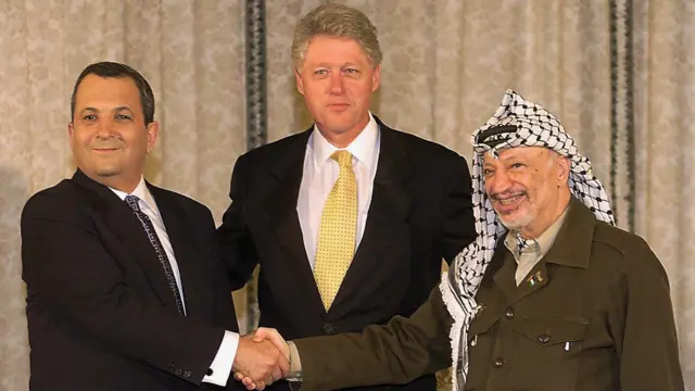 Ehud Barak y Yasir Arafat estrechan las manos sonrientes, con el presidente estadounidense Bill Clinton entre ambos.