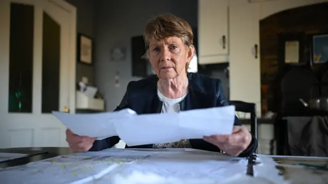Una mujer mayor está sentada a la mesa de la cocina. Sostiene una hoja de papel en cada mano. Sobre la mesa, frente a ella, hay más documentos y hojas de papel. Tiene el pelo corto, castaño y canoso, y viste una camiseta blanca y una chaqueta azul. Al fondo, hay armarios de cocina y una puerta de cocina con dos ventanas de cristal.
