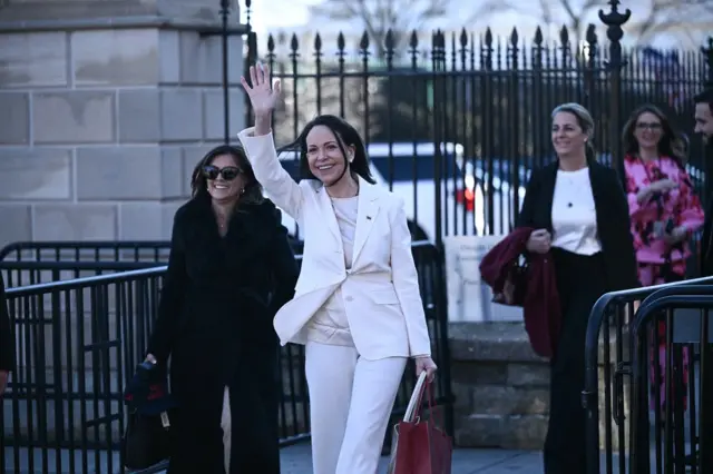 María Corina Machado , vestida de blanco y con una cartera marrón, saluda. A su lado hay una mujer con lentes de sol, vestida de negro y atrás, otra mujer con una blusa blanca y un traje negro