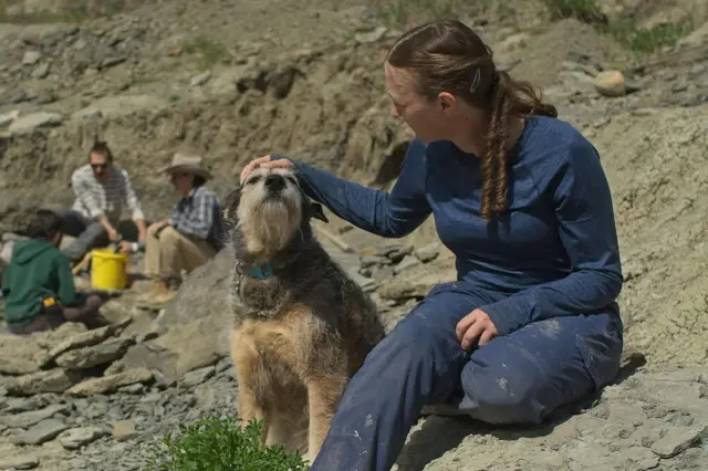 El perro de la profesora Bamforth, Aster, se sienta junto a ella sobre las rocas mientras acaricia la cabeza del can, con gente excavando en busca de fósiles al fondo.