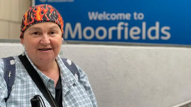 Sheila Irvine sonríe a la cámara junto a un cartel azul que da la bienvenida a Moorfields, el hospital oftalmológico londinense donde se la implantó la nueva teconología médica. 