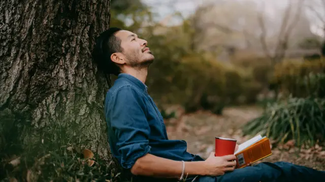 Un hombre sostiene un libro y un vaso en medio de un bosque. Tiene un rostro tranquilo 