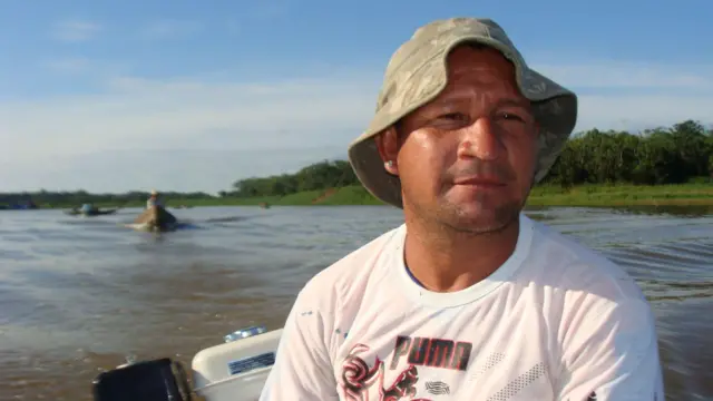 Pedro Canízio lleva una camiseta blanca y un sombrero de color caqui a bordo de su barco, con otros barcos y riberas verdes detrás de él.