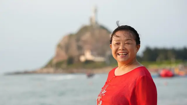 Una Su Min sonriente con una camiseta roja de pie junto al mar y con una colina borrosa en el fondo.