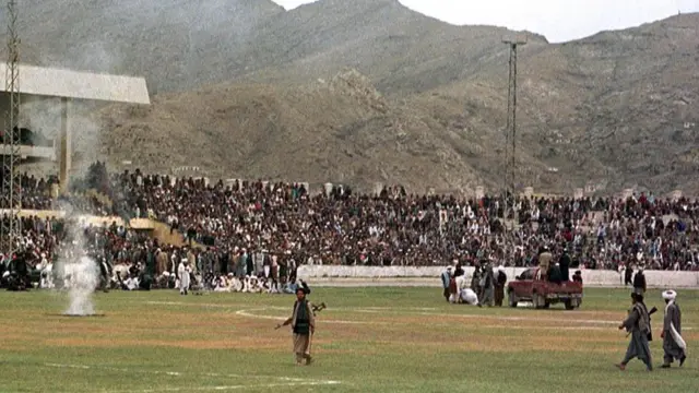 Un grupo de hombres con trajes tradicionales afganos portan armas mientras caminan por el césped en medio de un estadio. A la derecha, un hombre está encorvado en el suelo junto a un grupo de hombres y una camioneta. Una multitud observa desde las gradas.