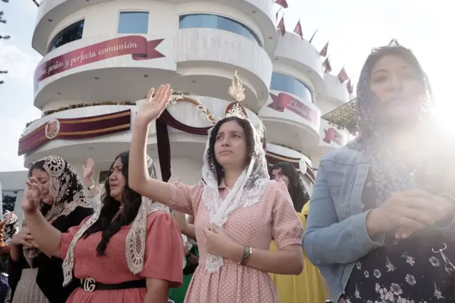 Mujeres feligresas de La Luz del Mundo, con velo en la cabeza, alzan la mano derecha mientras rezan durante la celebración llamada 
