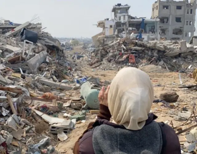 Una mujer con un velo de color ligero y un abrigo marrón está de espaldas a la cámara. Frente a ella están las ruinas de dos edificios destruidos y grandes pilas de escombros y tierra   