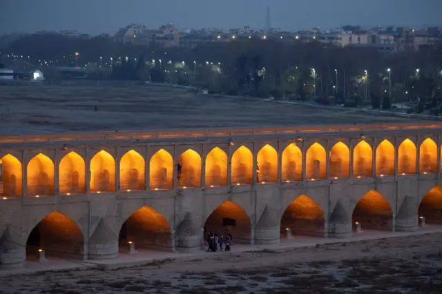 Iluminado al anochecer, el Puente de los 33 Arcos de principios del siglo XVII de la ciudad de Isfahán se extiende sobre el lecho seco del río Zayandeh Rud, mientras un grupo de peatones lo cruza.