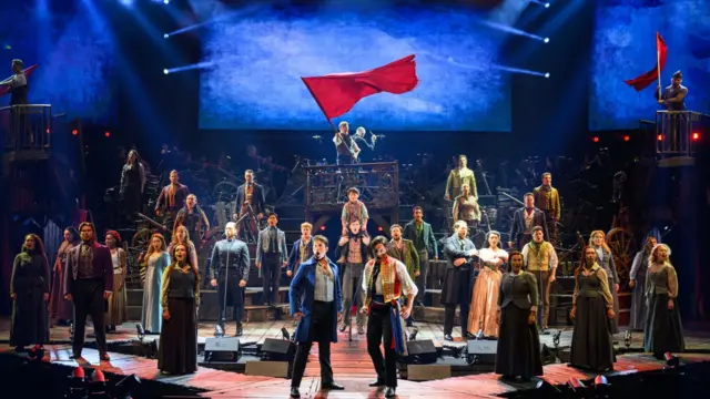 El elenco actuando durante la rueda de prensa de Los Miserables en Sidney, Australia. El elenco, vestido con trajes que recuerdan a la ropa de la época de la Revolución Francesa, se coloca en formación sobre el escenario. Al fondo, un hombre ondea una bandera roja y se encuentra de pie sobre una recreación de las barricadas que se utilizaban como defensa durante los combates.
