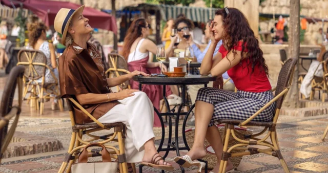 Dos mujeres ríen sentadas junto a dos copas de vino en la mesa de una terraza. 