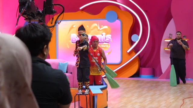 Rayyan Arkan Dikha, vestido con un traje tradicional malayo negro y gafas de sol, se sube a una silla y baila delante de una cámara.