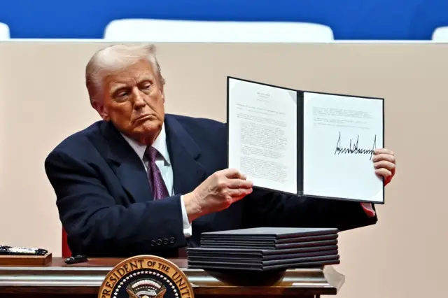 Trump, sentado frente a un escritorio sobre el que reposan varias carpetas, muestra al público una de ellas en las que se ve un documento firmado por él.