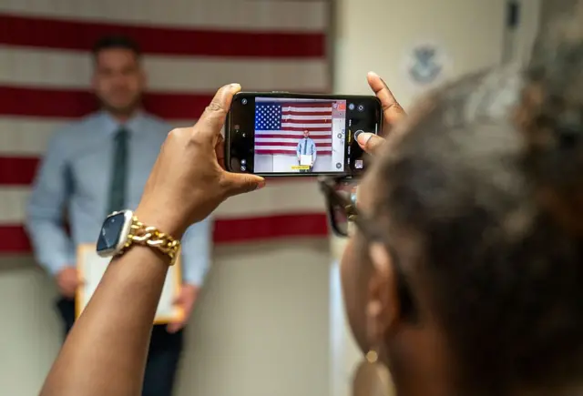 Un mujer toma una foto a un hombre frente a una bandera estadounidense en una ceremonia de naturalización de EE.UU.