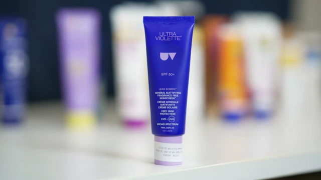 Un tubo de protector solar de color morado y azul se encuentra sobre un mostrador, con una variedad de otros protectores solares borrosos en el fondo.