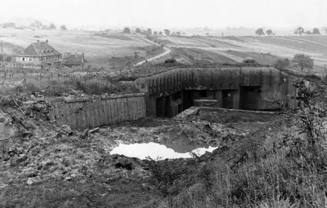 Un búnker de la Segunda Guerra Mundial destruido. Está en un campo abierto con una casona grande en el fondo