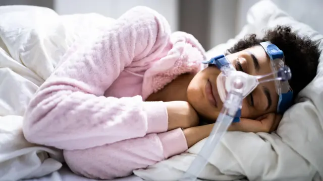 Una mujer joven duerme de lado en la cama con una máscara que cubre su nariz y está conectada a un largo tubo plástico