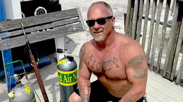 Johnny Mulford, un hombre de cabello gris corto, con una pequeña barba gris, está agachado sin camisa por lo que son visibles sus tatuajes. Lleva pantalones cortos y gafas de sol, y está arrodillado junto a unos cilindros que parecen contener aire y lo que parece ser un arpón.