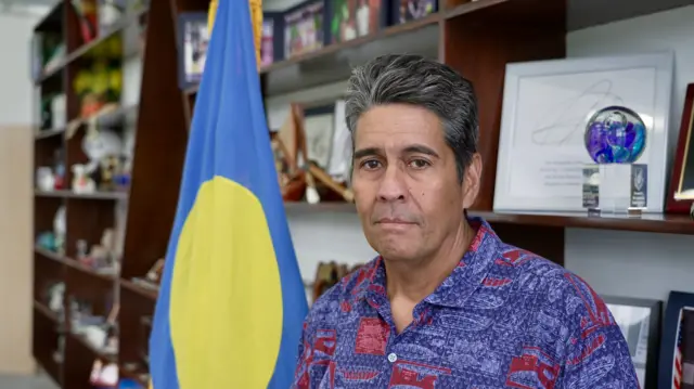 El presidente de Palaos, Surangel Whipps Jr., con una camisa azul y roja delante de la bandera del país.