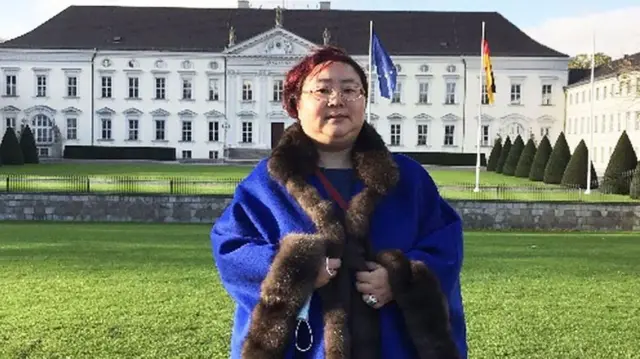 Una mujer con un abrigo de piel azul posa en un jardín frente a una mansión de color blanco.