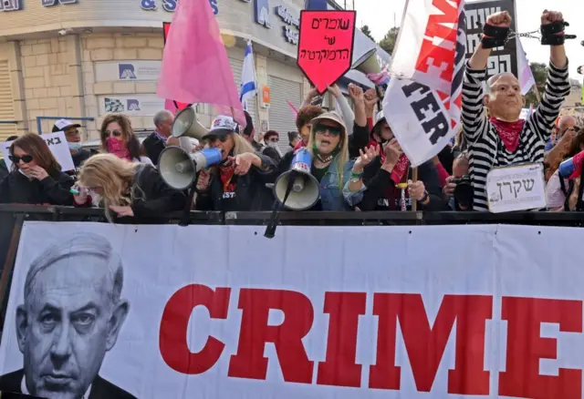 Manifestantes en Jerusalén con megáfonos y una pancarta que muestra una imagen de Netanyahu con la palabra 