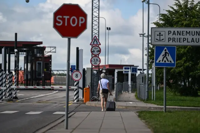 Una mujer con una maleta pequeña se acerca a un cruce de caminos. Hay una barrera a su izquierda y puertas y vallas frente a ella. Hay una señal de roja de Pare en primer plano.