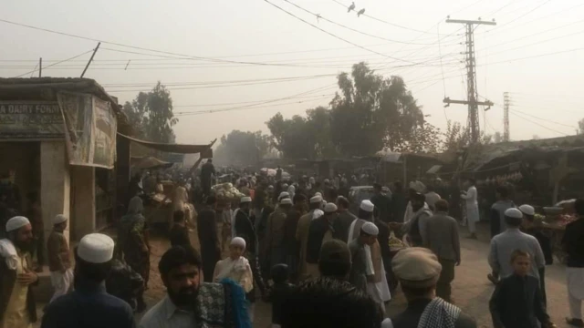 Una calle concurrida y polvorienta, llena de hombres, la mayoría con ropa tradicional afgana y sombreros sin ala. Hay edificios destartalados y torres de alta tensión que sostienen cables eléctricos sobre un cielo gris de fondo.