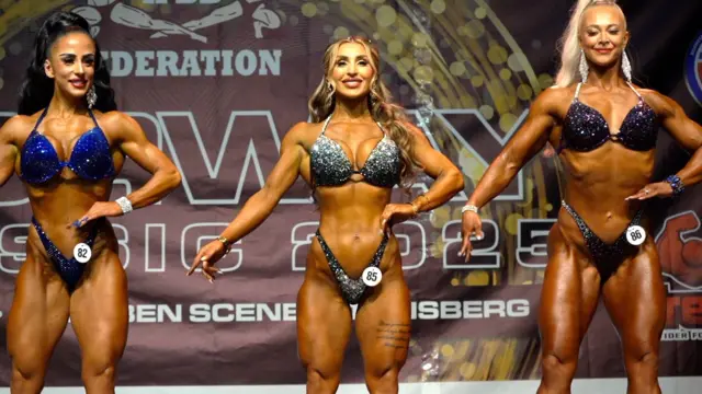 Tres mujeres bronceadas en trajes de baño brillantes, posan sonrientes mostrando su definición muscular sobre un escenario.