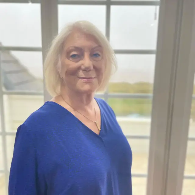 Carolyn porta una camisa azul y está parada frente a una ventana mirando a cámara   