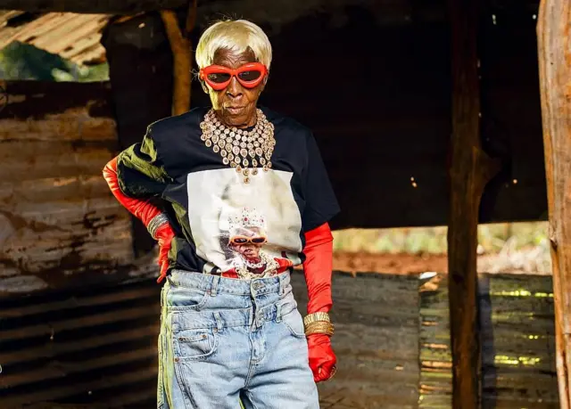 Margret Chola con jeans, un collar, guantes, gafas, una camiseta estampada con su imagen y una peluca rubia  