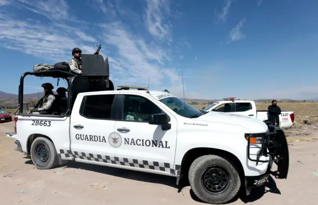 Dos autos de la Guardia Nacional de México, con varios efectivos ubicados en el remolque de uno de ellos y otro apostado junto a uno de ellos, en un paisaje desértico en México.
