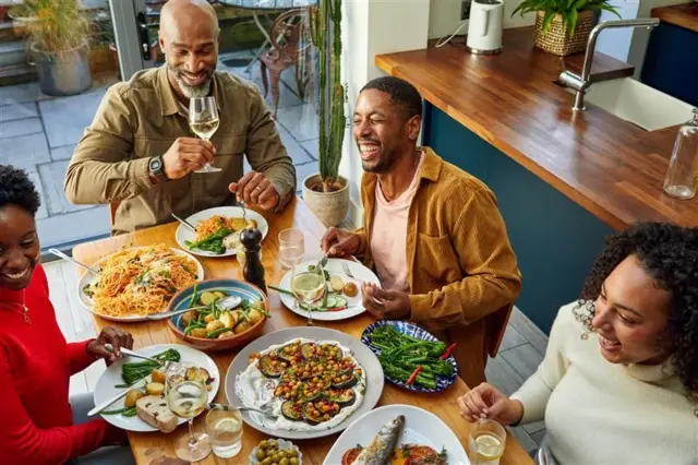 Una familia se ríe y divierte alrededor de una mesa con comida. Hay platos con brócoli, papas, aceitunas, pescado y berenjena