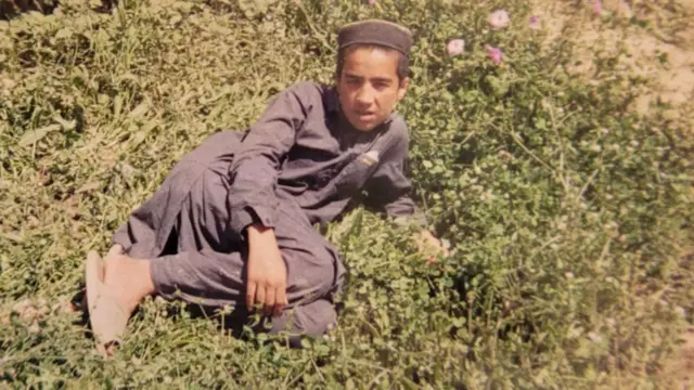 Fotografía a color de un adolescente tumbado de lado en un terreno cubierto de vegetación. Lleva una túnica tradicional afgana azul oscuro, pantalones, sandalias y un sombrero sin ala.
