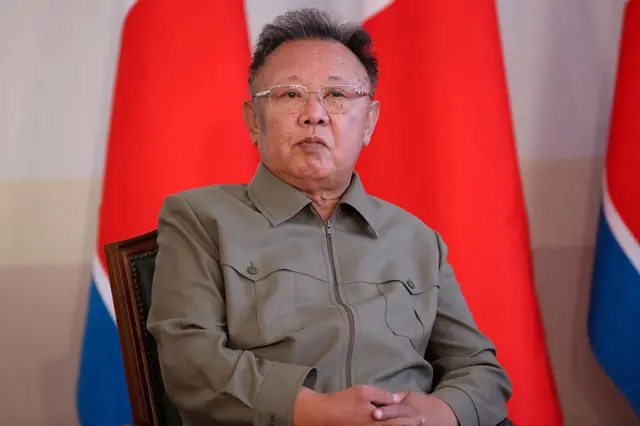El exlíder de Corea del Norte, Kim Jong Il, está sentado en una silla con una camisa verde y gafas, con banderas de Corea del Norte en el fondo.