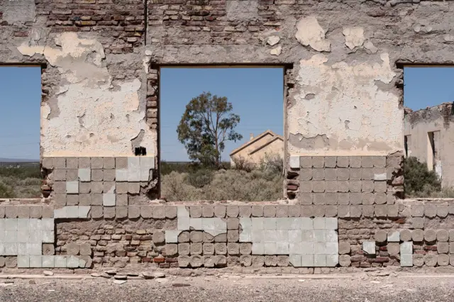 Una pared en ruinas a través de la cual se divisa una casa y un árbol