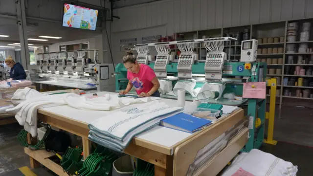 Una trabajadora con una camiseta rosa está elaborando toallas en la fábrica Matouk, en Fall River, Massachusetts. Está examinando una toalla blanca sobre una gran mesa de trabajo, de pie frente a una gran máquina de color verde azulado que parece estar bordando diseños sobre otras toallas.